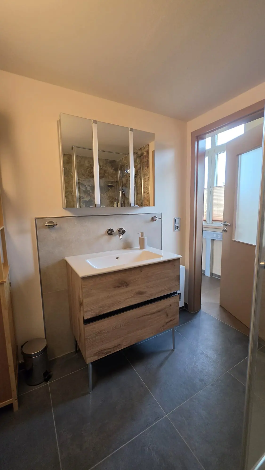 Badezimmer der Ferienwohnung Zahna-Loft — Waschtisch mit Spiegelschrank auf grauen Bodenfliesen, daneben die offene Tür zum hellen Nebenraum