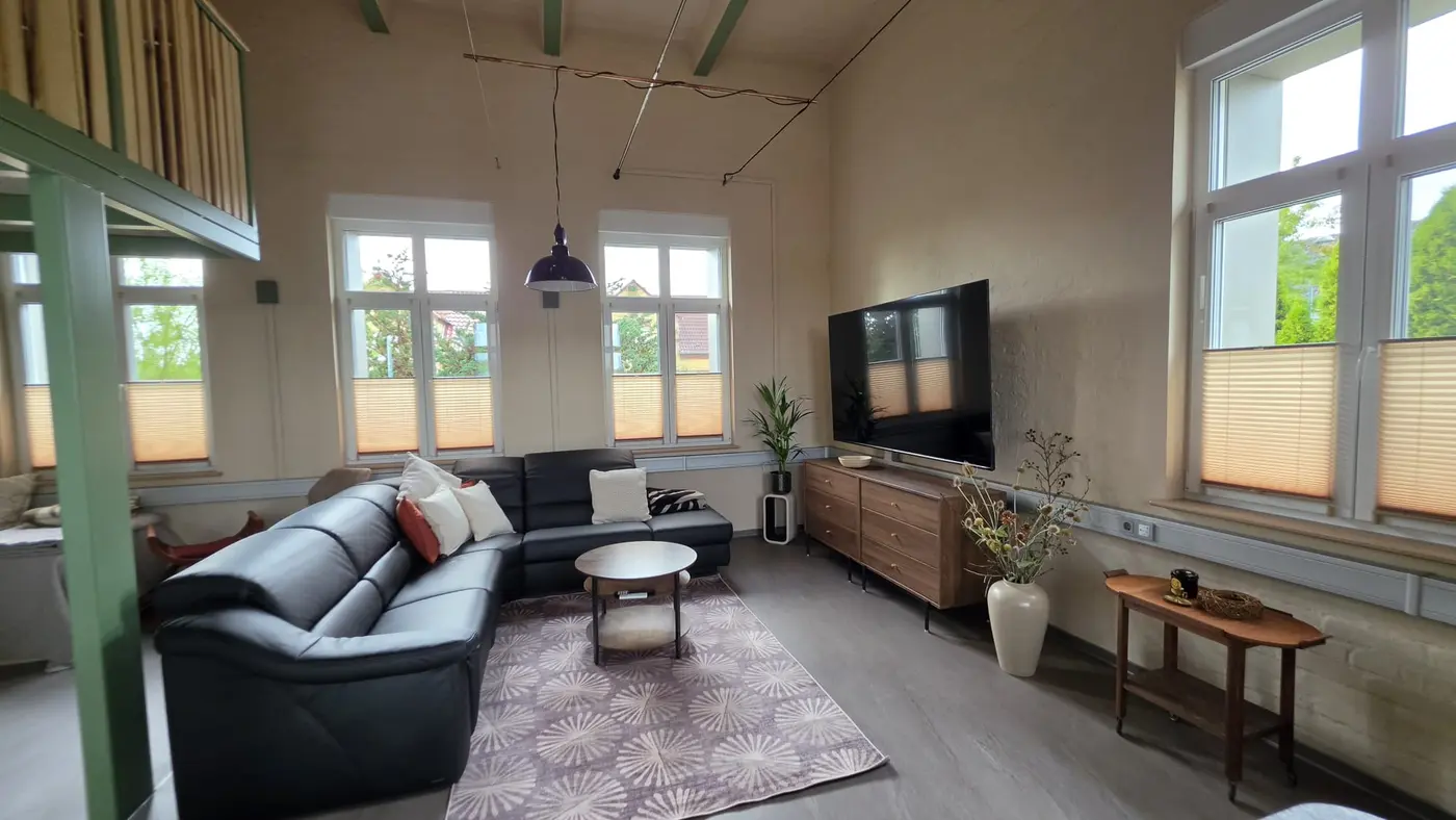 Offener Wohn- und Essbereich der Ferienwohnung Zahna-Loft — Ledersofa mit rundem Couchtisch, Sideboard und Fernseher vor dem Fensterband