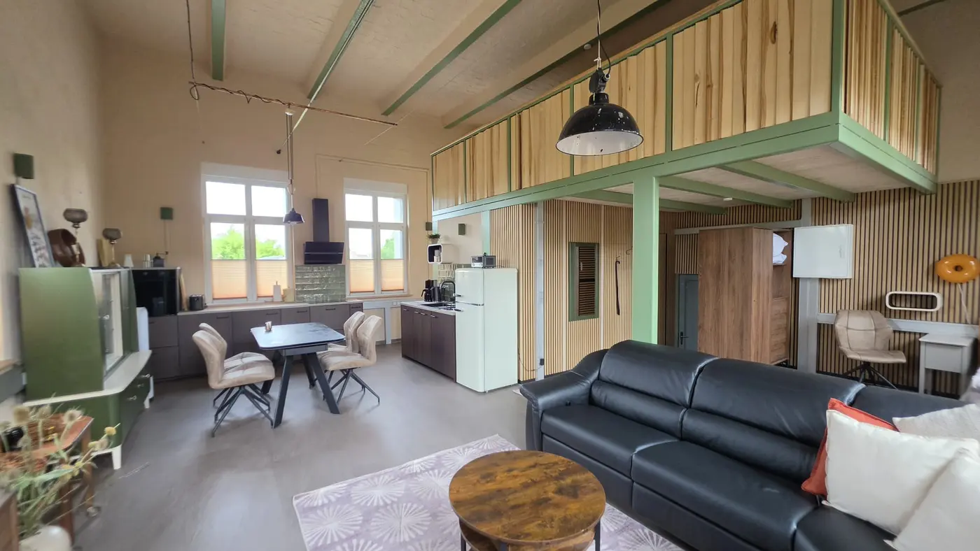 Offener Wohn- und Essbereich der Ferienwohnung Zahna-Loft — Gesamtansicht mit grünen Stahlträgern, Esstisch, Küche und Ledersofa