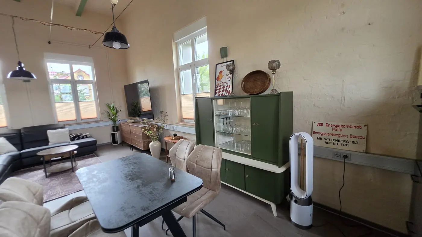 Offener Wohn- und Essbereich der Ferienwohnung Zahna-Loft — Esstisch und grüne Vintage-Vitrine unter den schwarzen Werkstattlampen, im Hintergrund das Ledersofa